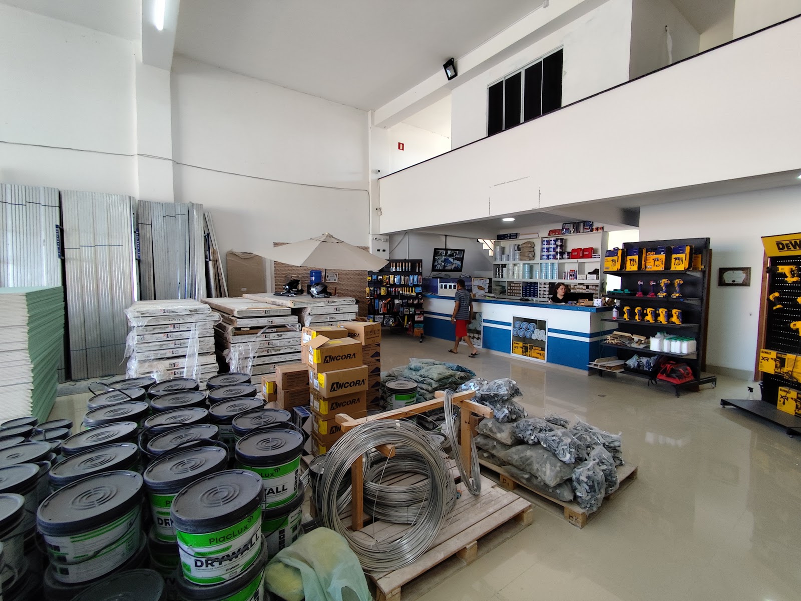 Salão da loja com baldes de massa para drywall, rolos de arame, placas empilhadas, balcão azul e expositores de ferramentas